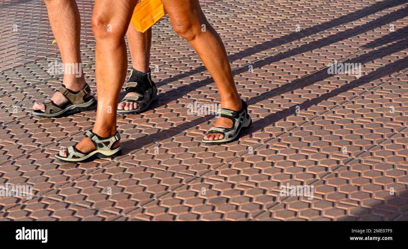 Gebräunte Beine von zwei Personen, die Sandalen tragen und auf gefliestem Pflaster laufen Stockfoto