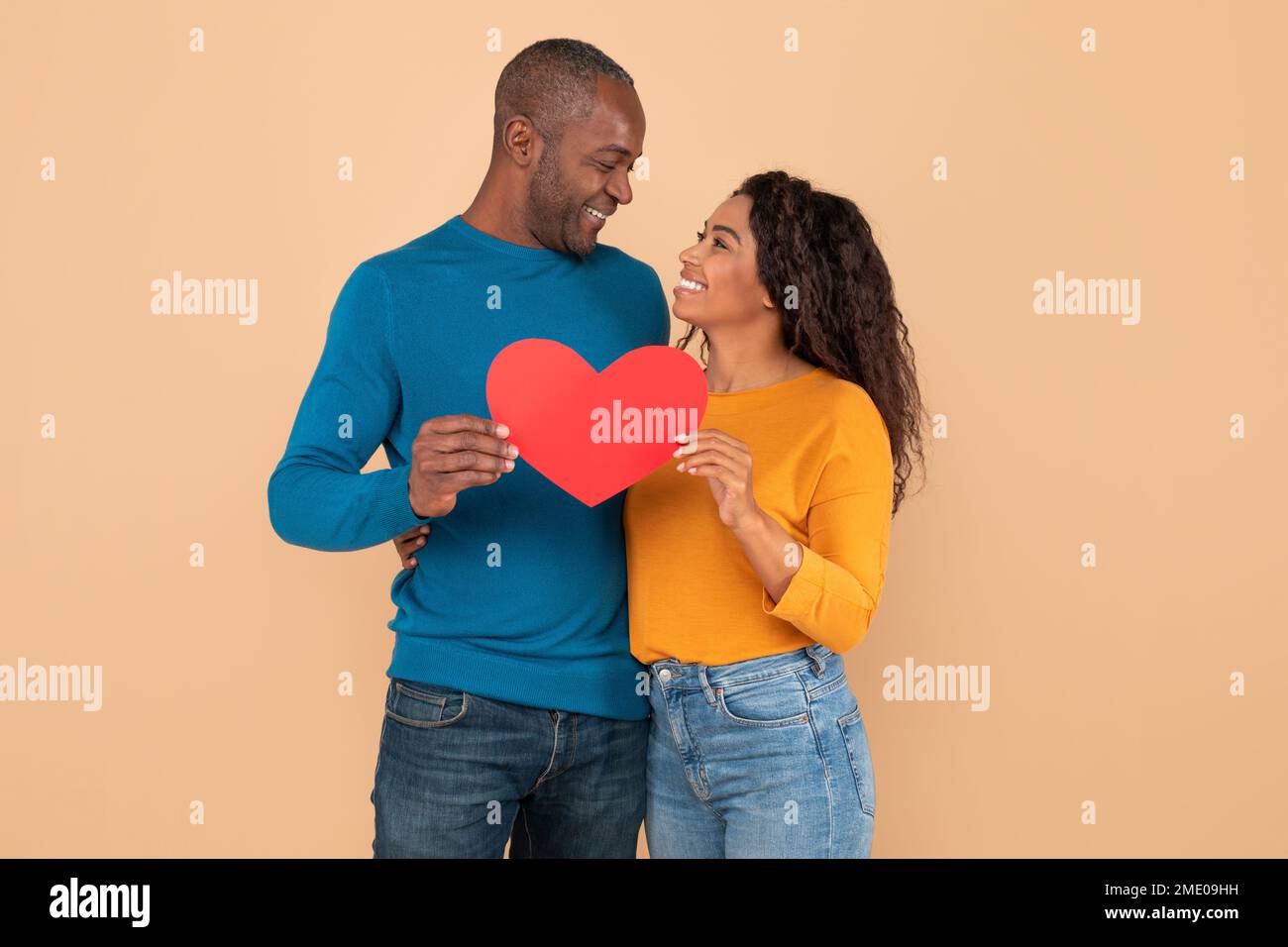 Valentinstag. Schwarzer Mann mittleren Alters und junge Frau umarmt, mit rotem Herzen, pfirsichfarbener Studiohintergrund Stockfoto