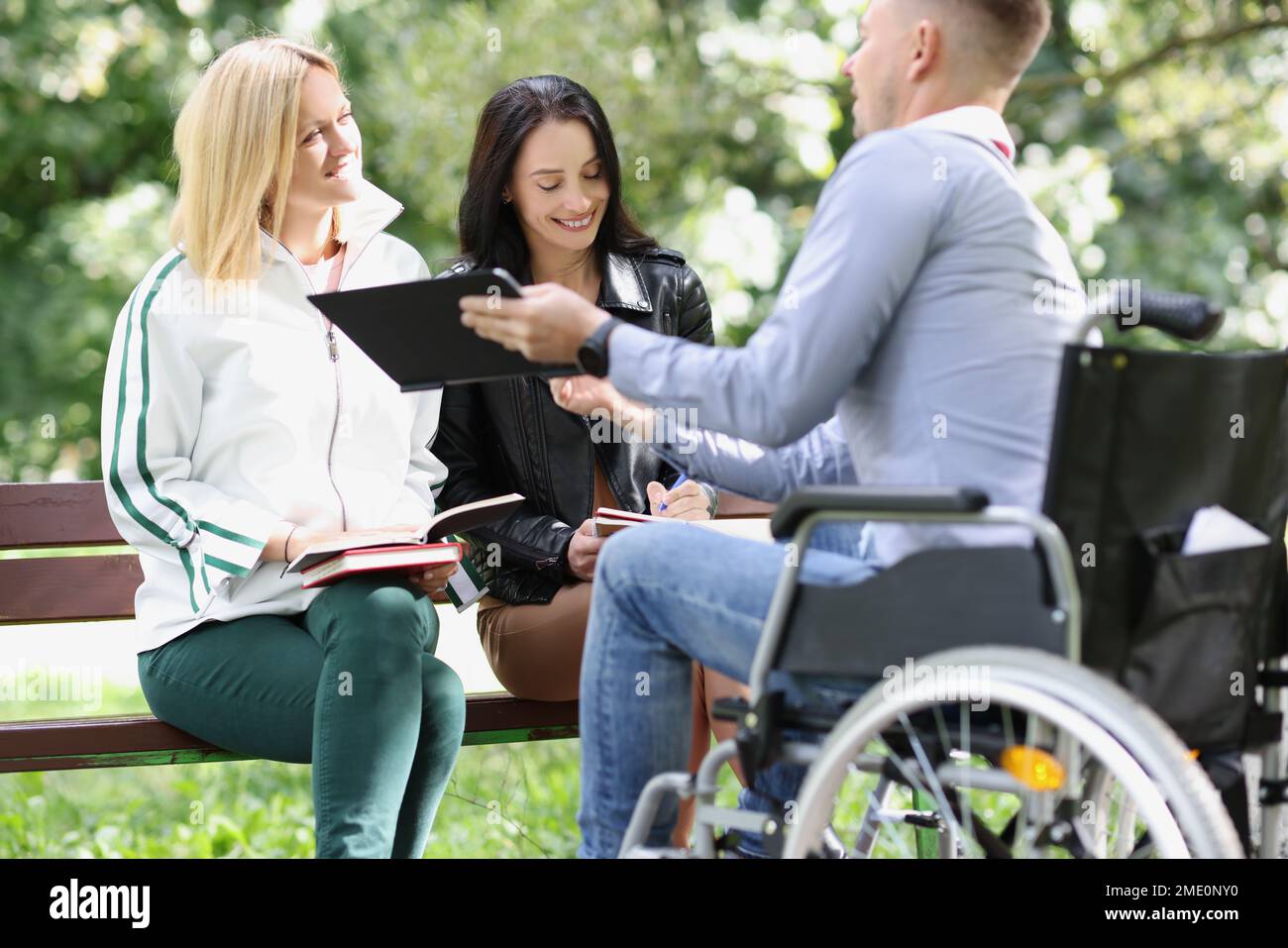 Zwei Studentinnen und ein männlicher Student studieren zusammen im Park, der Mann sitzt im Rollstuhl. Stockfoto