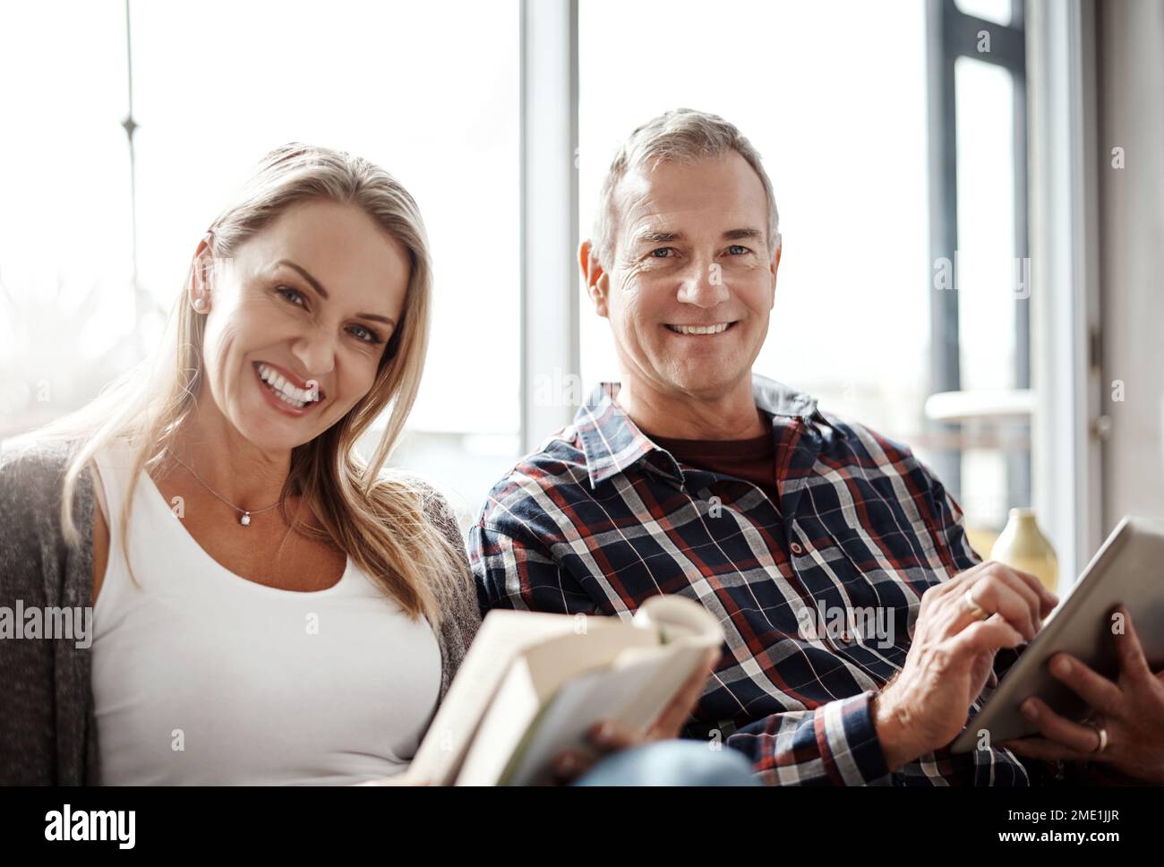 So sehen unsere Sonntage aus. Eine reife Frau liest ein Buch, während ihr Mann zu Hause ein digitales Tablet auf dem Sofa benutzt. Stockfoto
