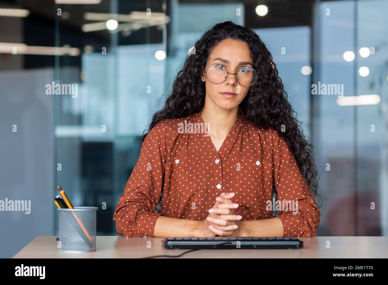 Porträt einer jungen Lehrerin aus Latino. Sie sitzt ernsthaft am Tisch im Büro, faltete ihre Hände auf den Tisch, blickt in die Kamera. Stockfoto