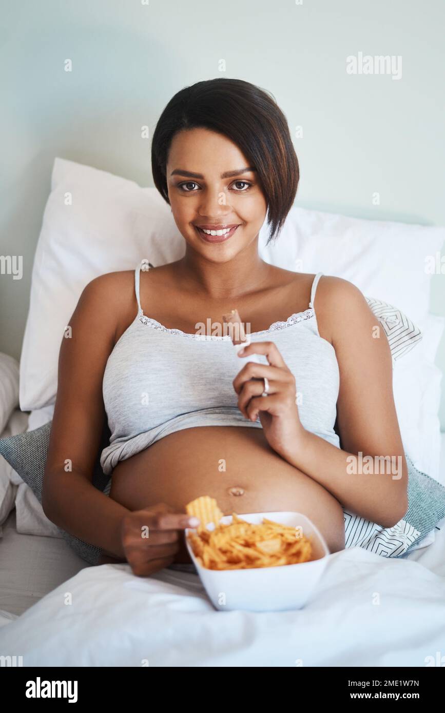 Das Baby will, was es will. Porträt einer attraktiven jungen schwangeren Frau, die Kartoffelchips und Schokolade im Bett isst. Stockfoto