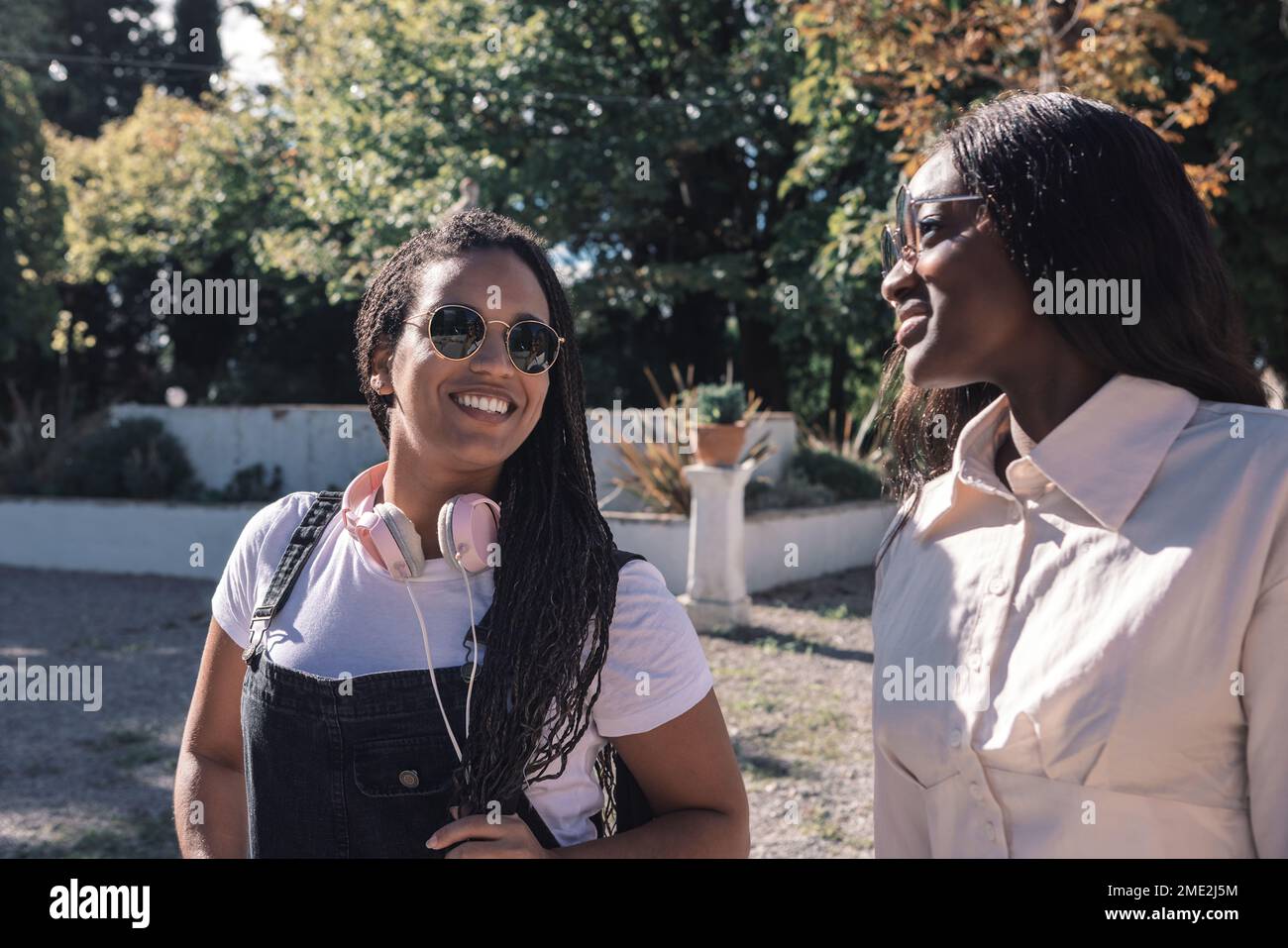 Lächelnde junge afroamerikanische Freundinnen in lässiger Kleidung und Sonnenbrille, die im Park neben grünen Bäumen stehen und sich gegenseitig umarmen Stockfoto