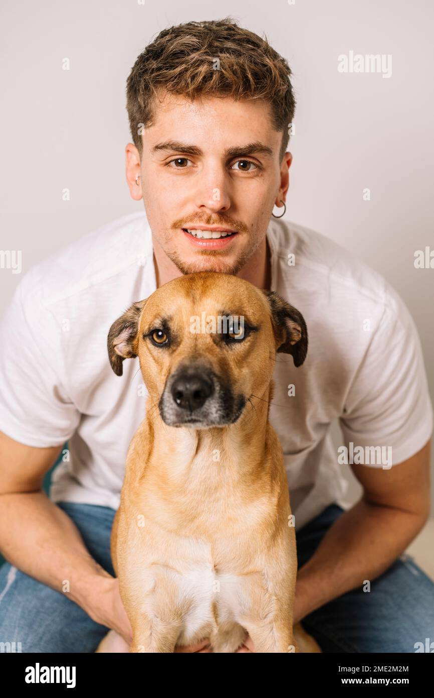 Lächelnder männlicher Besitzer in weißem Hemd und Jeans, der hinter einem niedlichen reinrassigen Hund sitzt und kuschelt, während er in die Kamera schaut Stockfoto