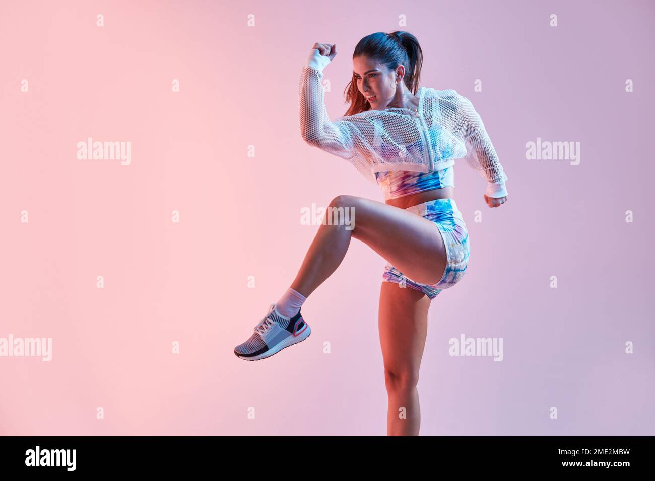 Blick auf eine fit junge Frau mit farbenfrohem Oberteil und Shorts, die hoch springen, mit erhobenem Bein und Arm an der rosa Wand im Studio Stockfoto