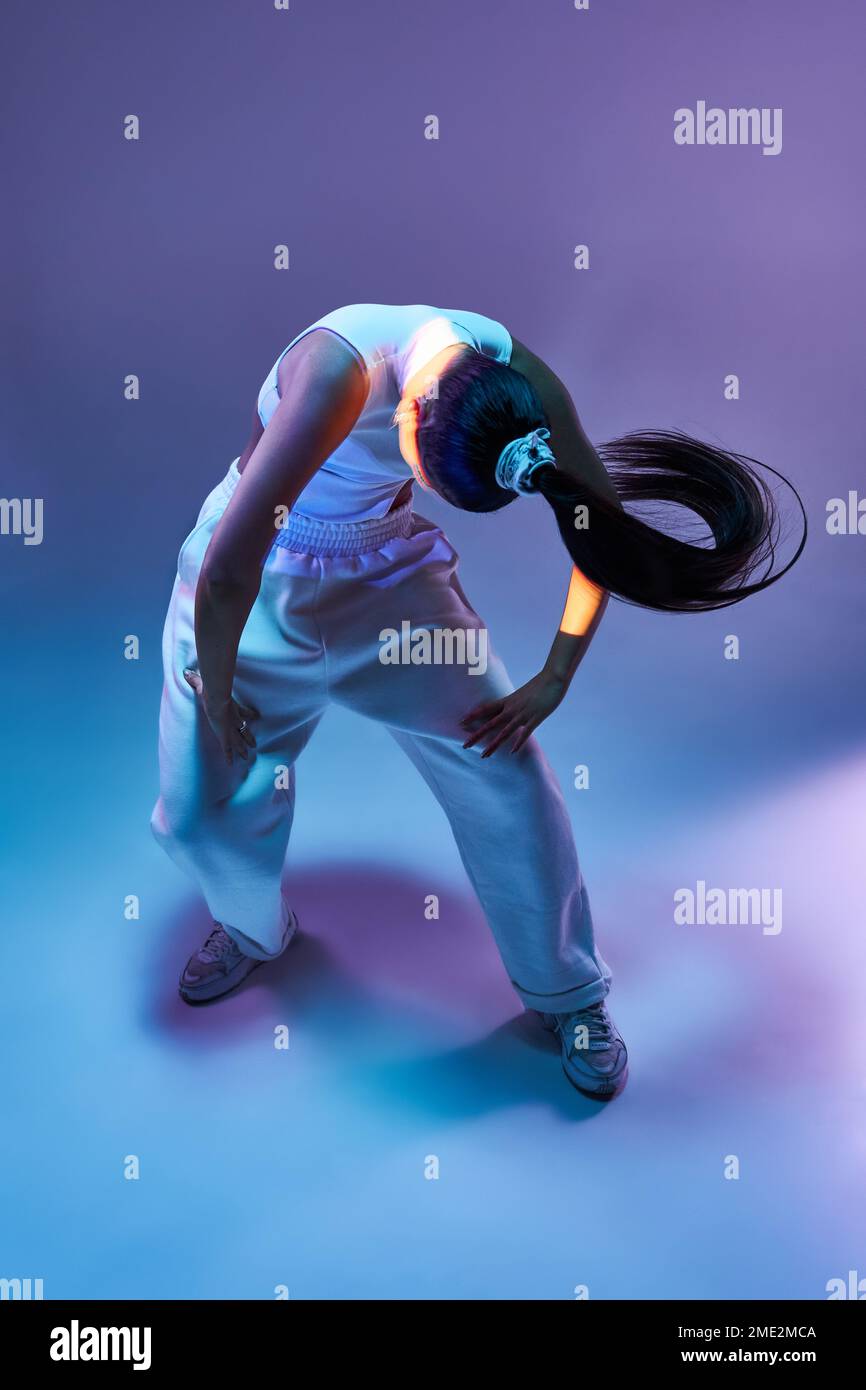 Die junge Tänzerin in sportlichen Hosen lehnt sich nach vorne und spielt Musik gegen violettes Neonlicht Stockfoto