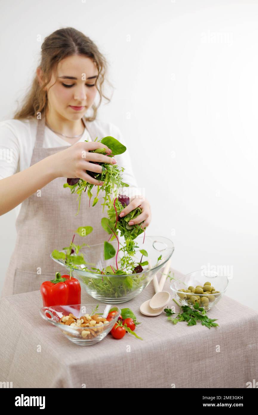 Das Mädchen mischt Salat mit den Händen Sie gießt ihn in eine Glasplatte vegetarisches Essen gesundes Essen Holzlöffel Schürze Tischdecke junge Frau Teenager bereitet Abendessen für die Familie zu Frühstück Mittagessen Stockfoto