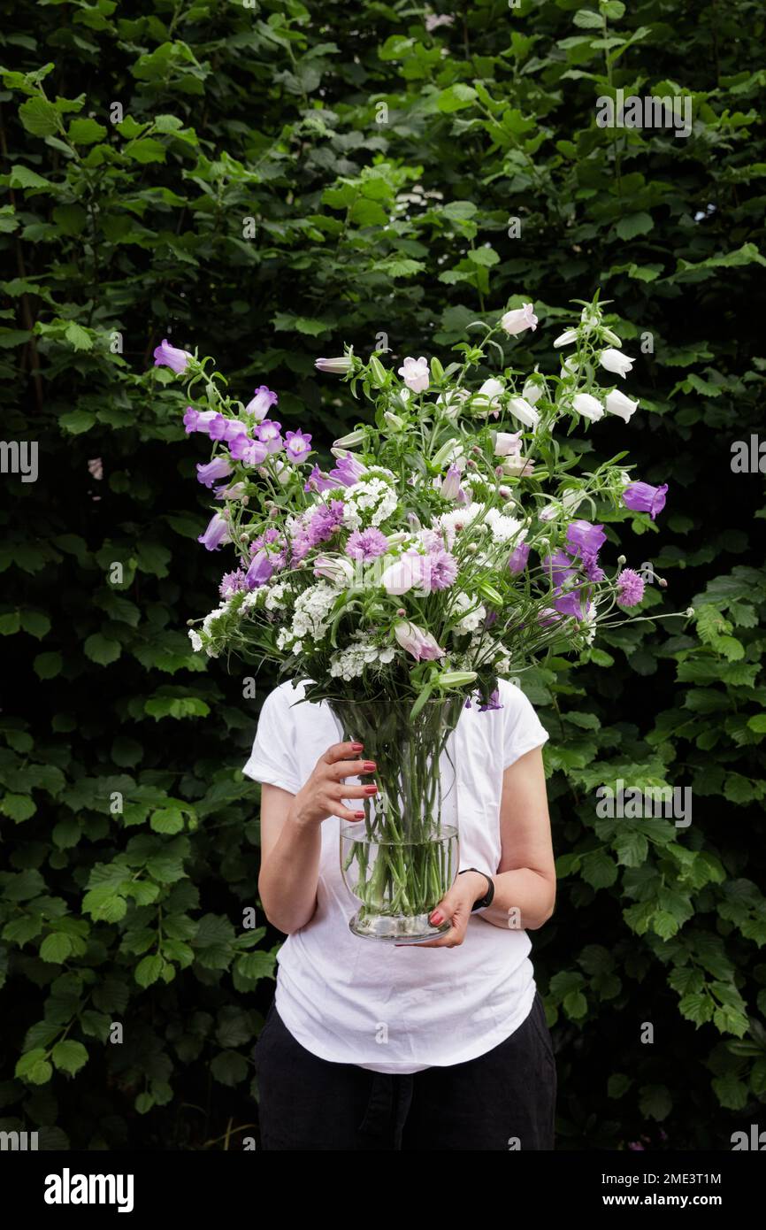 Reife Frau, die das Gesicht mit canterbury-Glockenblumen im Glasglas verdeckt Stockfoto