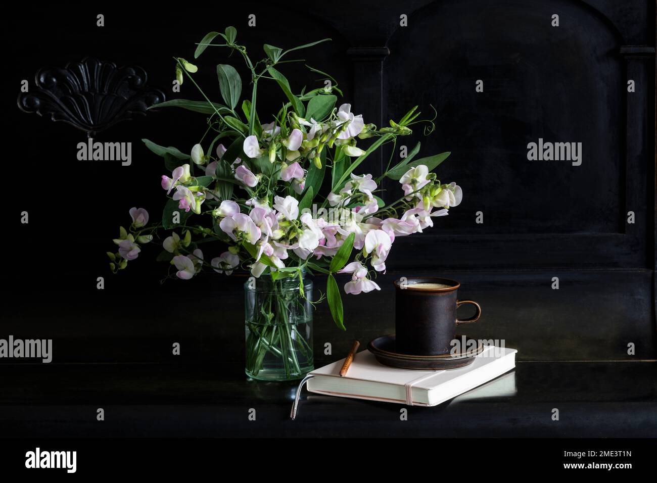 Sweetpea-Blumen im Glasgefäß bei Kaffeetasse und Tagebuch auf schwarzem Klavier Stockfoto