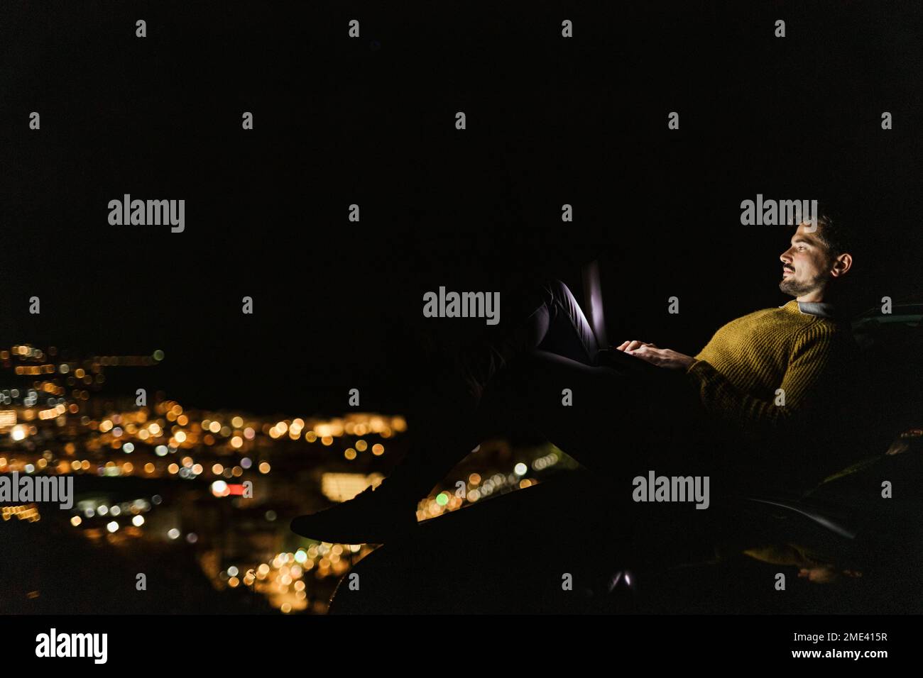 Mann, der nachts auf einem Hügel über der beleuchteten Stadt sitzt und ein Notebook benutzt Stockfoto