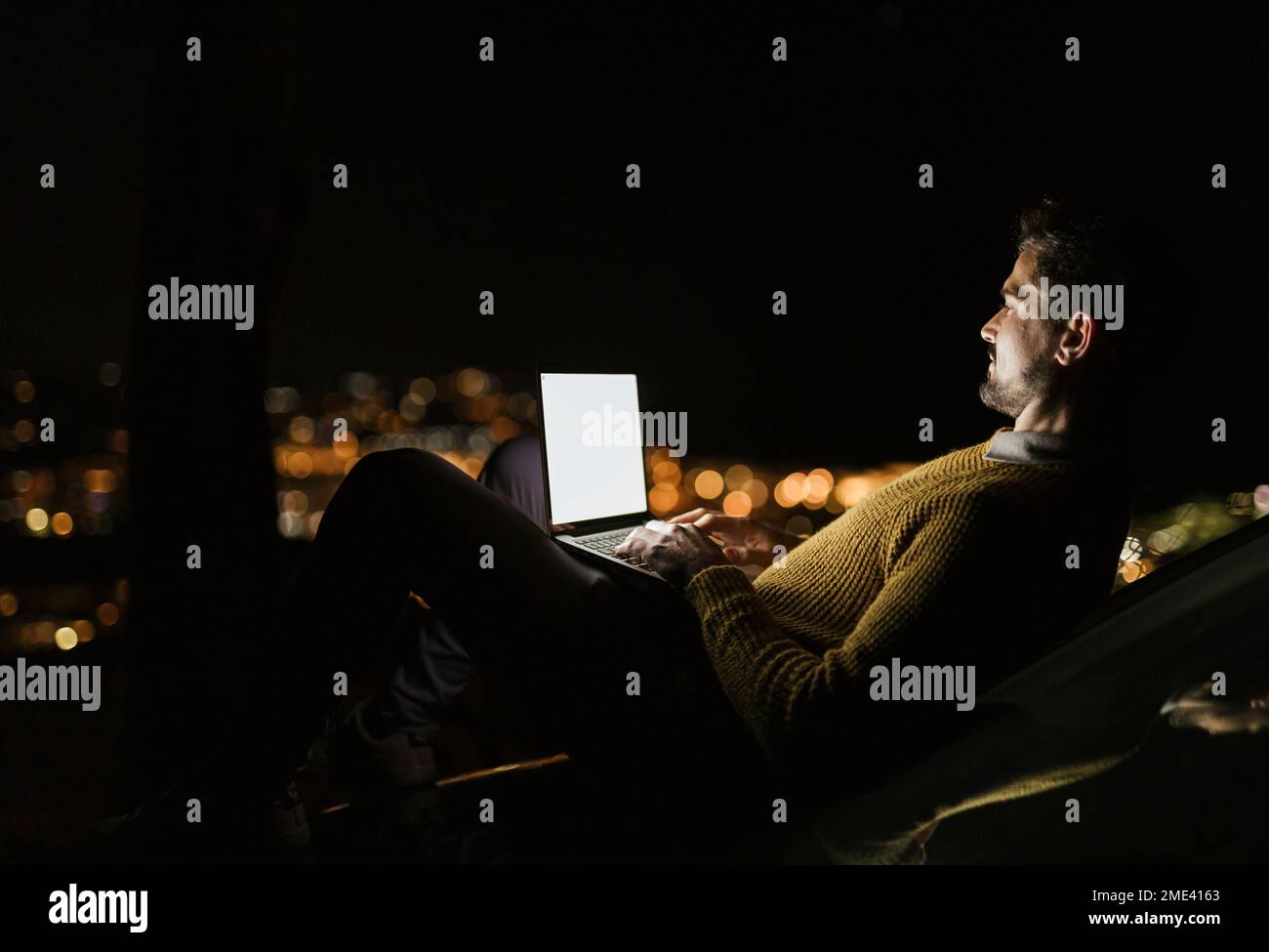 Mann, der nachts auf einem Hügel über der beleuchteten Stadt sitzt und ein Notebook benutzt Stockfoto