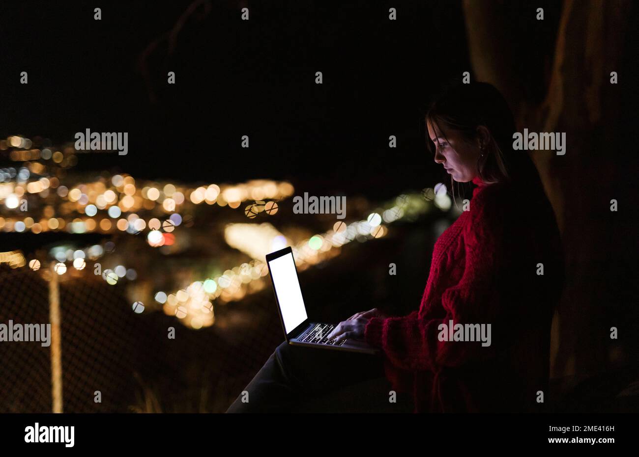 Frau, die nachts auf einem Hügel über einer beleuchteten Stadt sitzt und ein Notebook benutzt Stockfoto