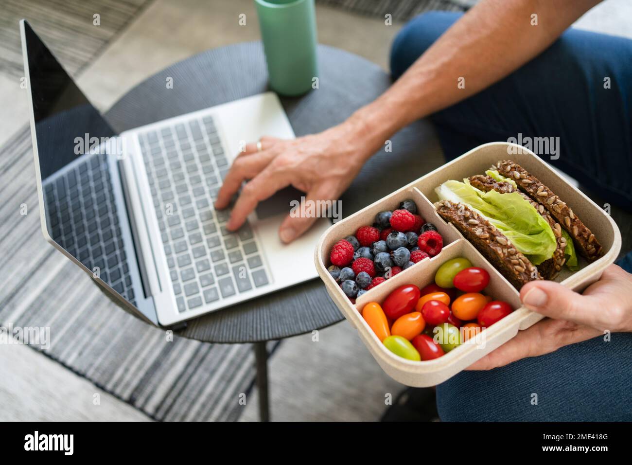 Hand eines Mannes, der eine Lunchbox mit gesundem Essen hält und zu Hause einen Laptop benutzt Stockfoto
