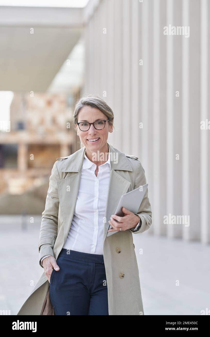 Selbstbewusste Geschäftsfrau, die den Tablet-PC im Stehen hält und die Hand in die Taschen steckt Stockfoto