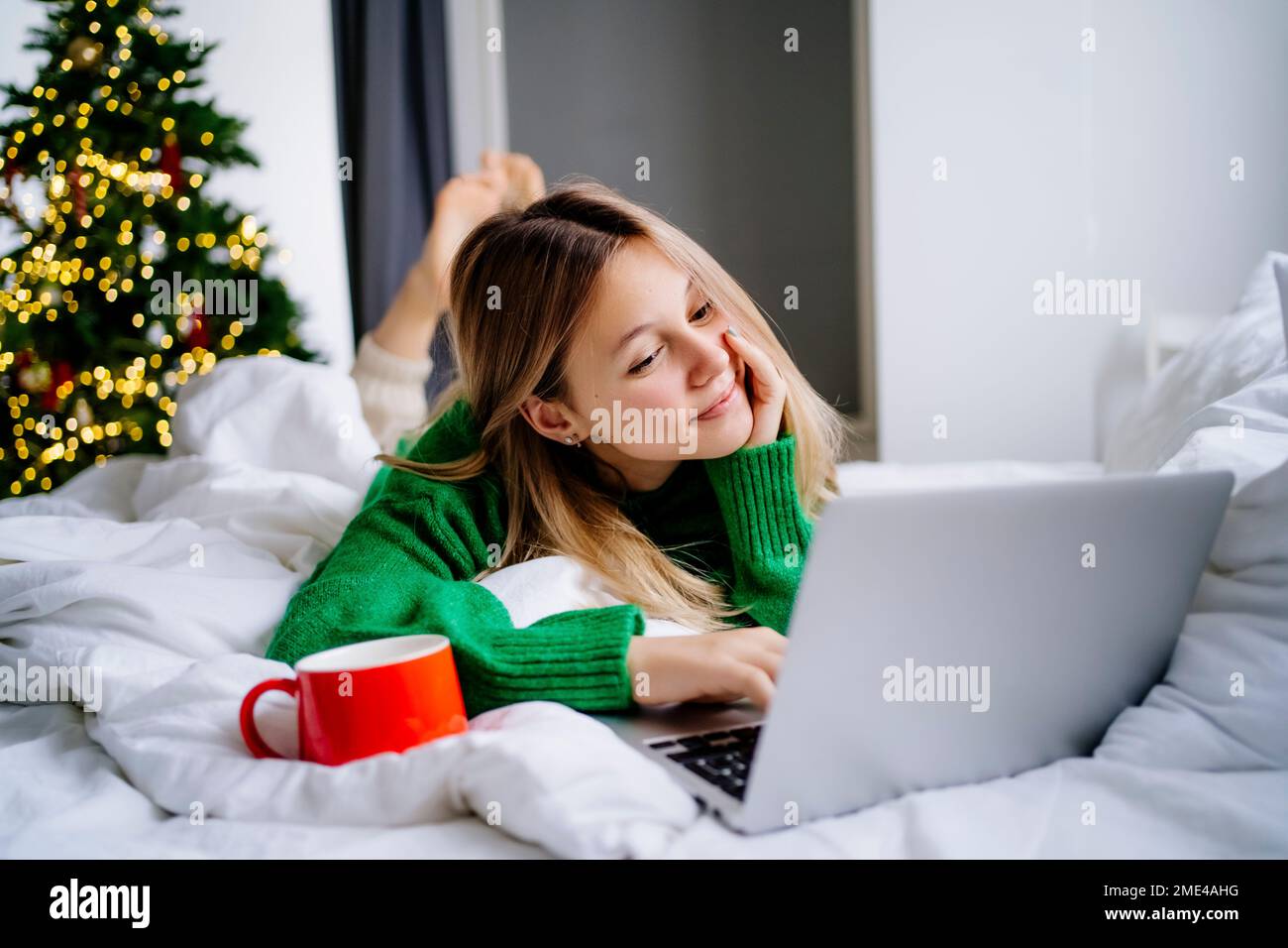 Lächelndes Mädchen, das sich zu Hause einen Film auf dem Laptop ansieht, mit roter Tasse auf dem Bett Stockfoto
