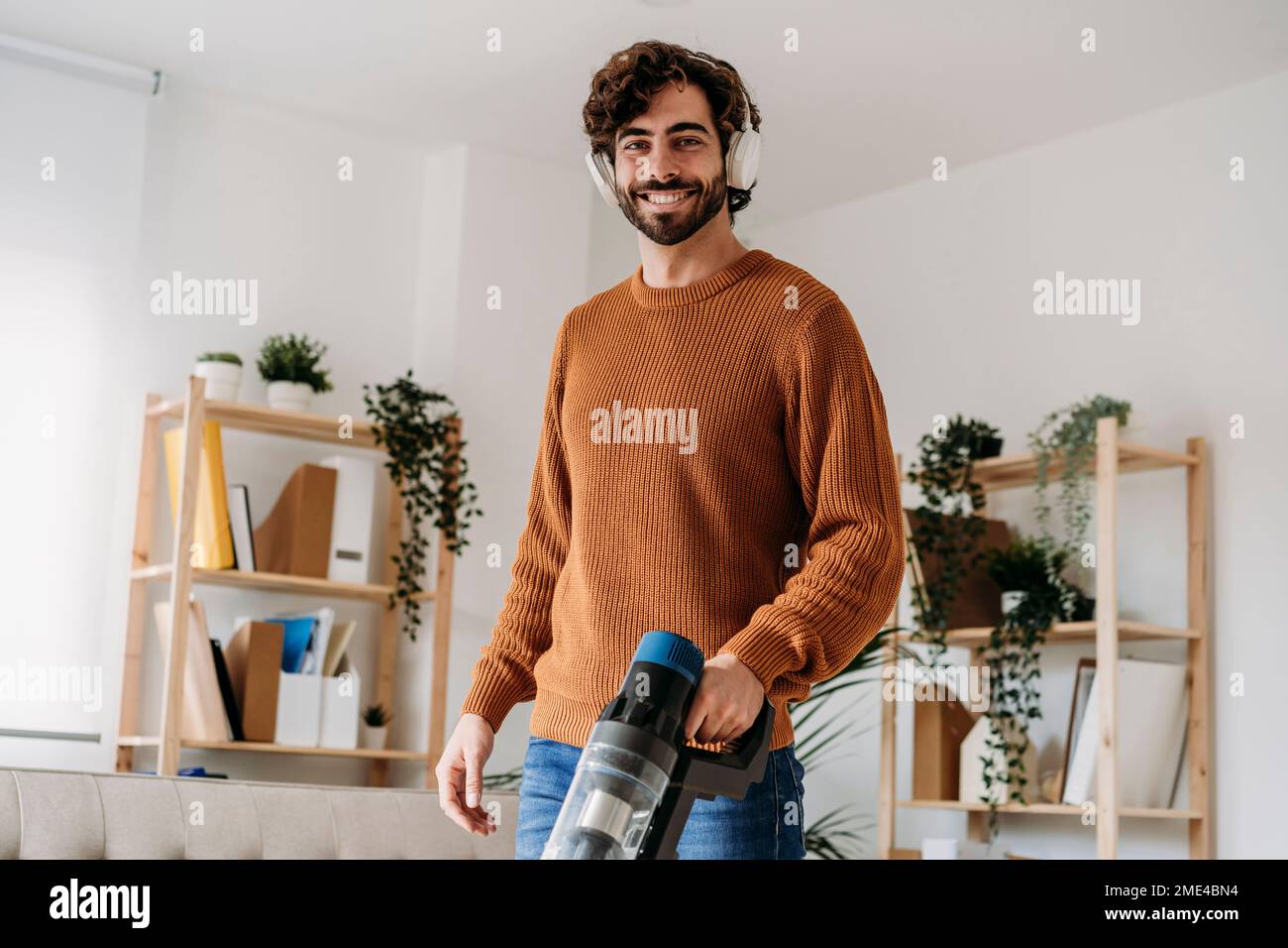 Lächelnder Mann mit kabellosen Kopfhörern, der sein Zuhause mit einem Staubsauger reinigt Stockfoto