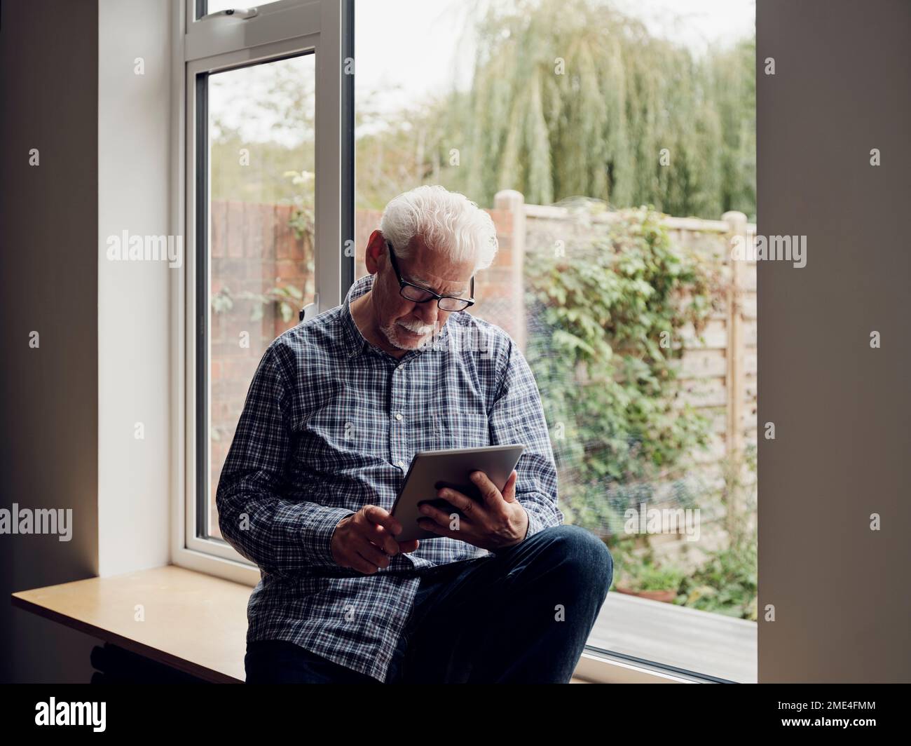 Seniorenmann am Fenster zu Hause mit einem digitalen Tablet Stockfoto