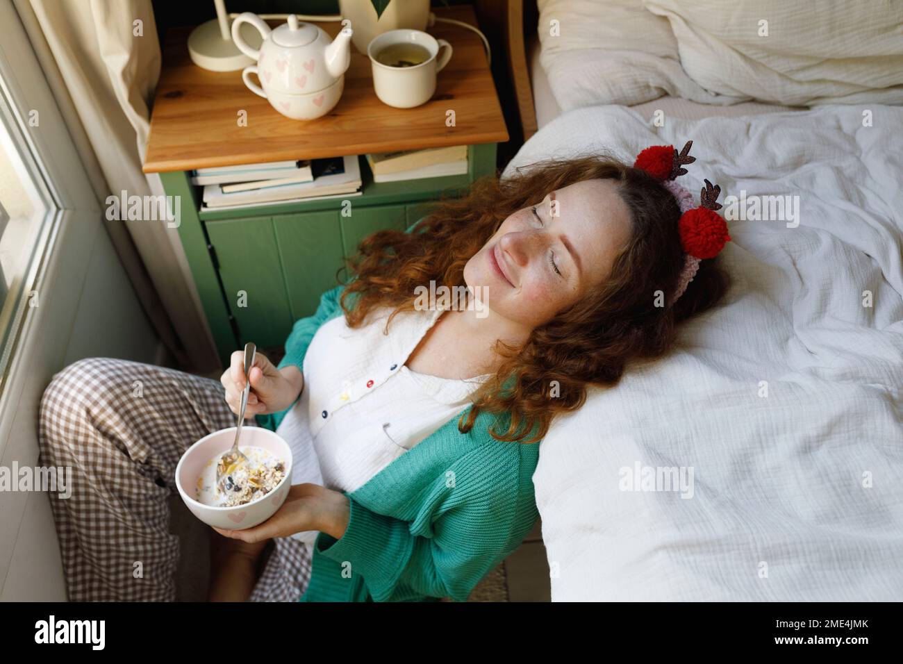 Lächelnde Frau mit geschlossenen Augen, die zu Hause eine Müslischale am Bett hält Stockfoto