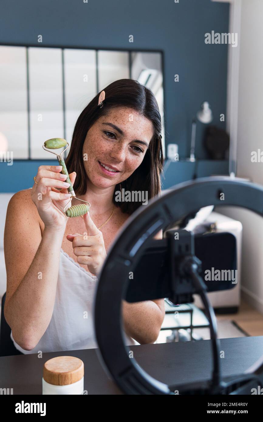 Lächelnde Frau, die zu Hause Jade-Stein-Roller auf dem Smartphone zeigt Stockfoto