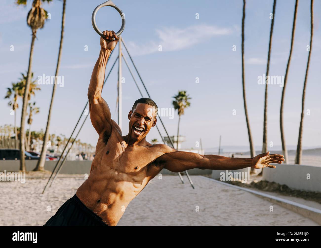 Fröhlicher Sportler, der sich am Strand an Turnringen festhält Stockfoto