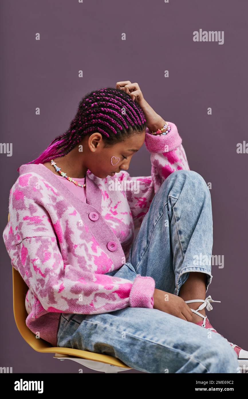 Vertikales seitliches Porträt eines schwarzen Mädchens im Teenageralter, das trendige Freizeitkleidung trägt und mit erhobenen Füßen auf dem Stuhl sitzt Stockfoto