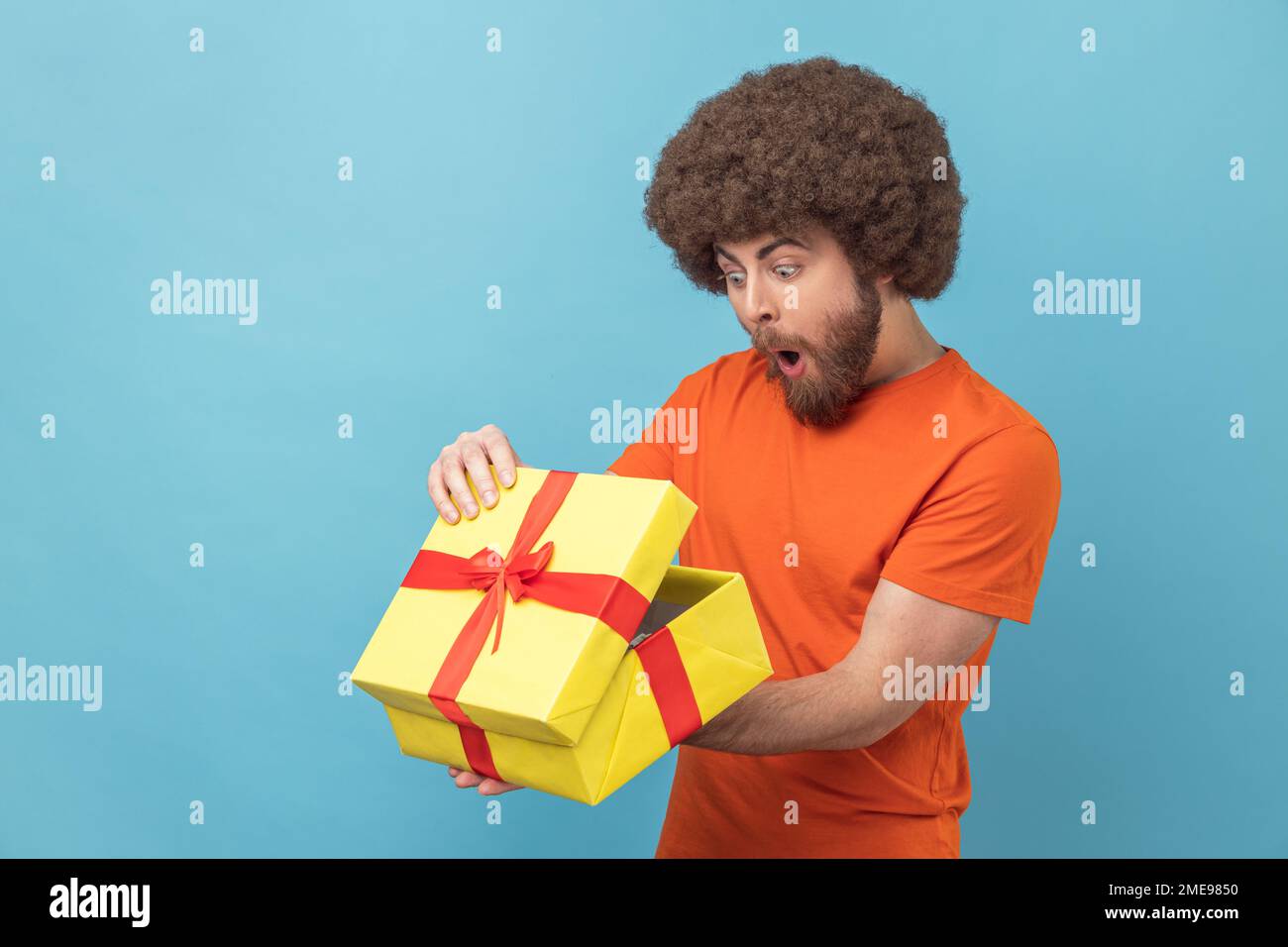 Porträt eines Mannes mit Afro-Haarschnitt, der ein orangefarbenes T-Shirt trägt, das Geschenk auspackt, in eine Schachtel mit schockiertem und verängstigtem Gesichtsausdruck schaut, Geburtstagsgeschenk. Innenstudio, isoliert auf blauem Hintergrund. Stockfoto
