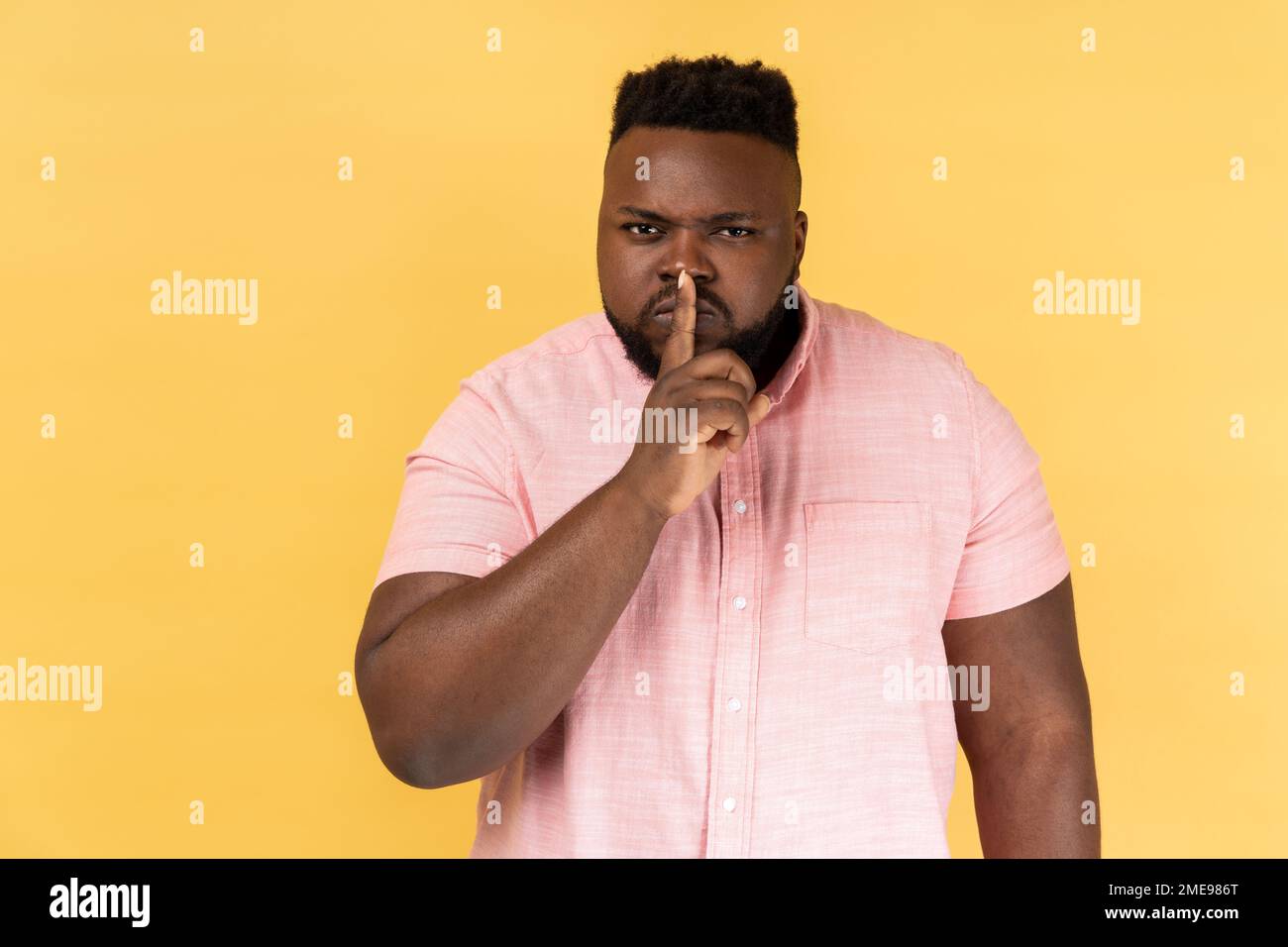 Schweigen Sie bitte. Portrait eines ernsthaften, strengen Mannes, der ein rosa Hemd trägt, steht, stille Geste zeigt, still sagt, geheim halten. Innenstudio-Aufnahme isoliert auf gelbem Hintergrund. Stockfoto