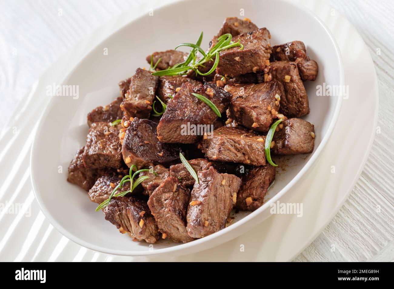 Nahaufnahme von gebratenen saftigen New york Strip Steak Bissen mit Butter, Rosmarin und Knoblauch in Schüssel auf weißem Holztisch Stockfoto