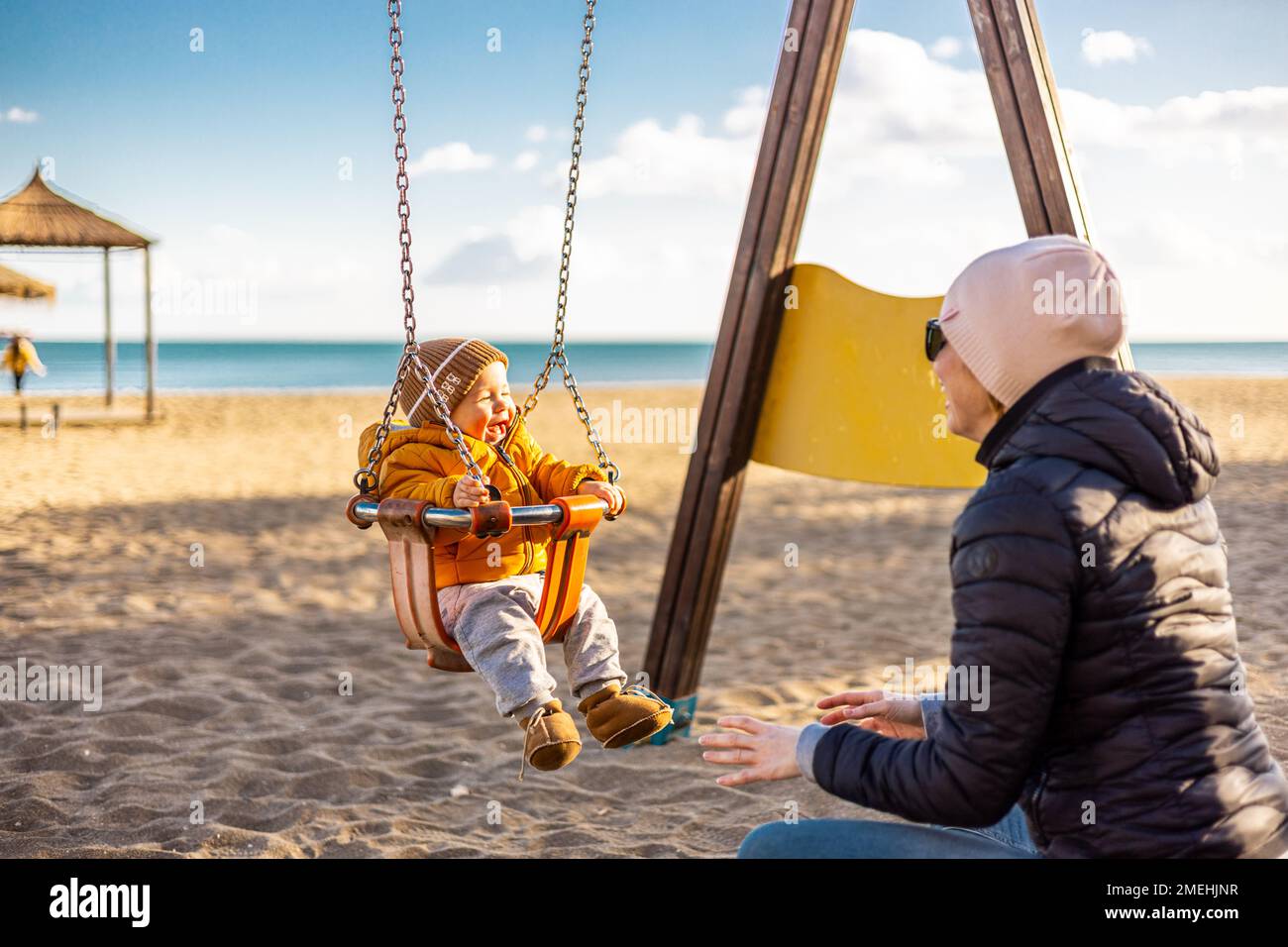 Mutter schubst ihr kleines Kind auf einer Schaukel auf dem Sandstrand im Freien an einem schönen sonnigen, kalten Wintertag in Malaga, Spanien Stockfoto
