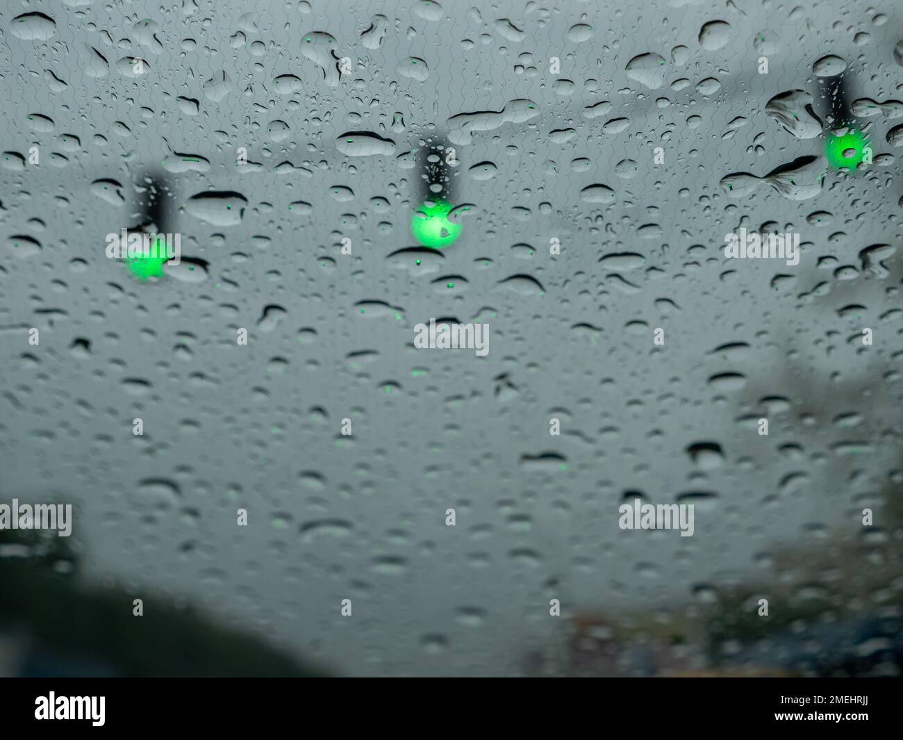 Nahaufnahme von Regentropfen auf einer Windschutzscheibe, verschwommene Ampeln mit grünen Signalen, die den Verkehr lenken. Ansicht vom Fahrersitz aus. Stockfoto