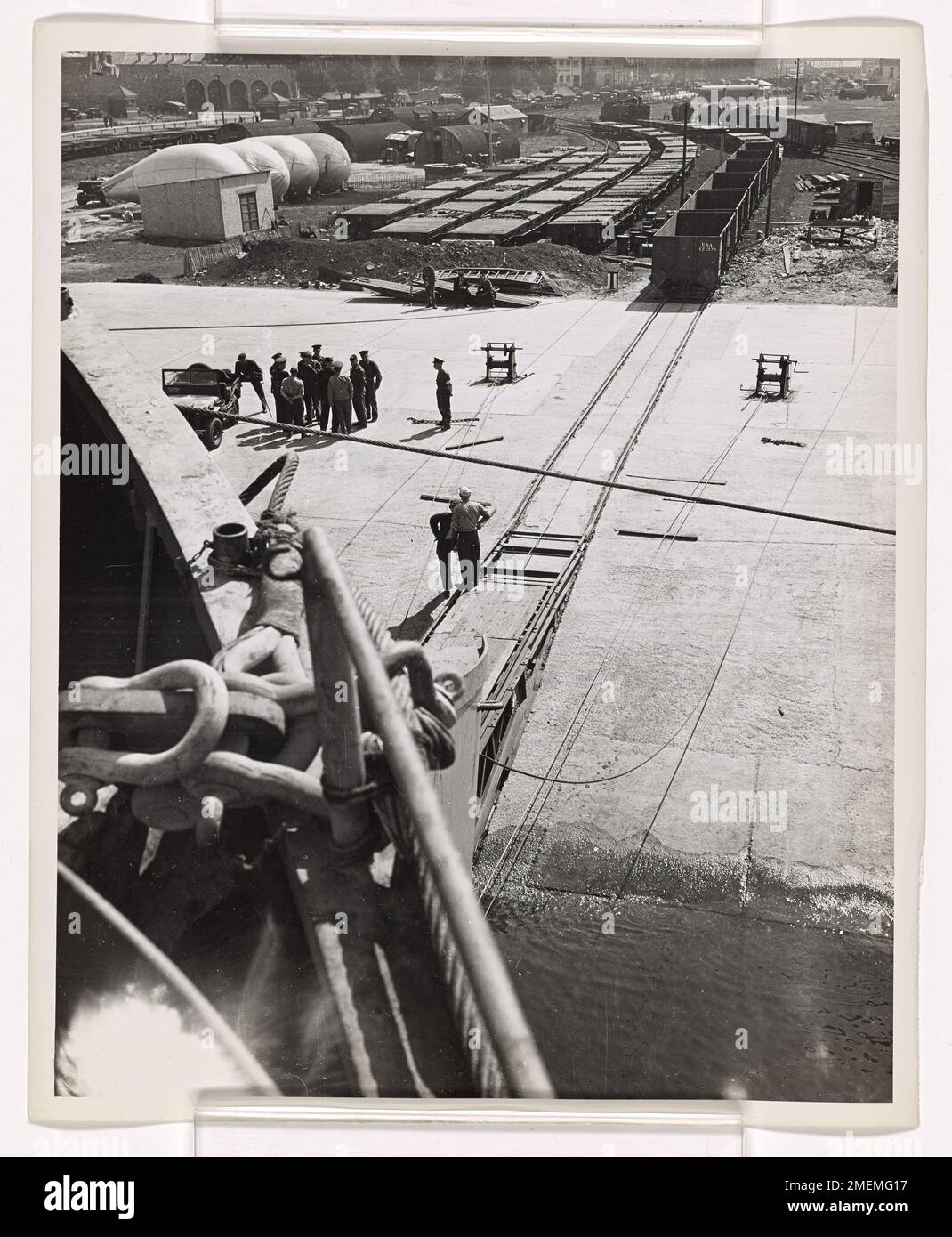 In einem britischen Hafen bereitet sich ein von der Küstenwache bemanntes LST (Landing Ship, Tank) auf die Schiene vor, wo Güterwagen des Army Transportation Corps auf das schienenbestückte Deck des Schiffs gerollt werden. Diese Wagen werden nach Frankreich transportiert und auf eine Landstraße geschoben, die sich bis zum Wasserrand erstreckt, wodurch die Verbindung zwischen den Eisenbahnstrecken in England und Frankreich für die militärische Logistik während des Zweiten Weltkriegs erleichtert wird. Stockfoto