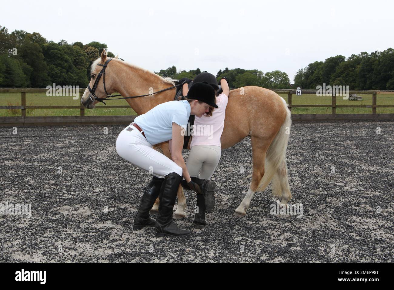 Lehrerin, die dem jungen Mädchen ein Bein hochmacht, während sie das palomino-Pony während des Reitstages in der Koppel besteigt Stockfoto