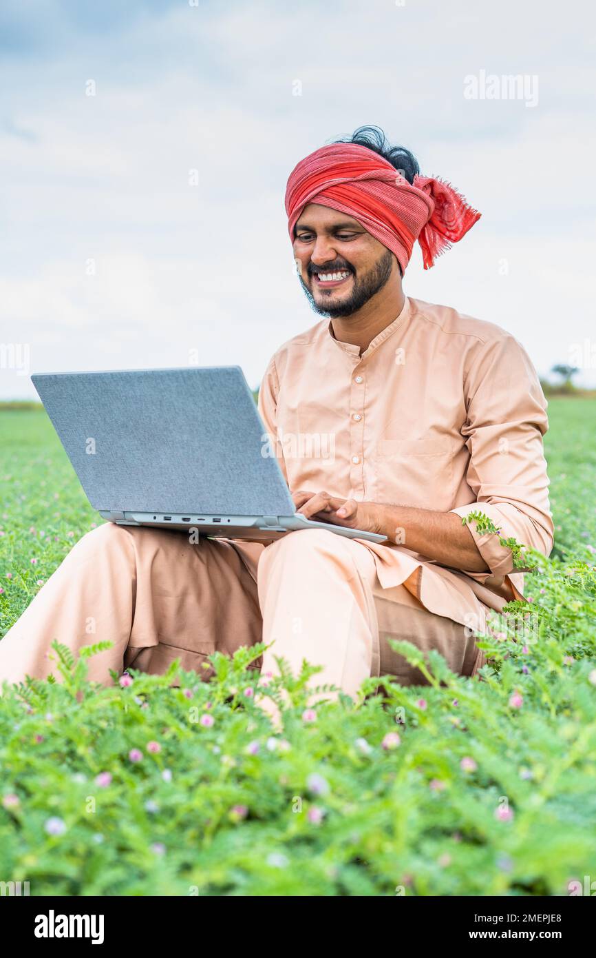 Vertikale Aufnahme eines fröhlichen, lächelnden jungen Landwirts, der auf dem Ackerland mit einem Laptop arbeitet – Konzept von Technologie, moderner Landwirtschaft und Entwicklung Stockfoto