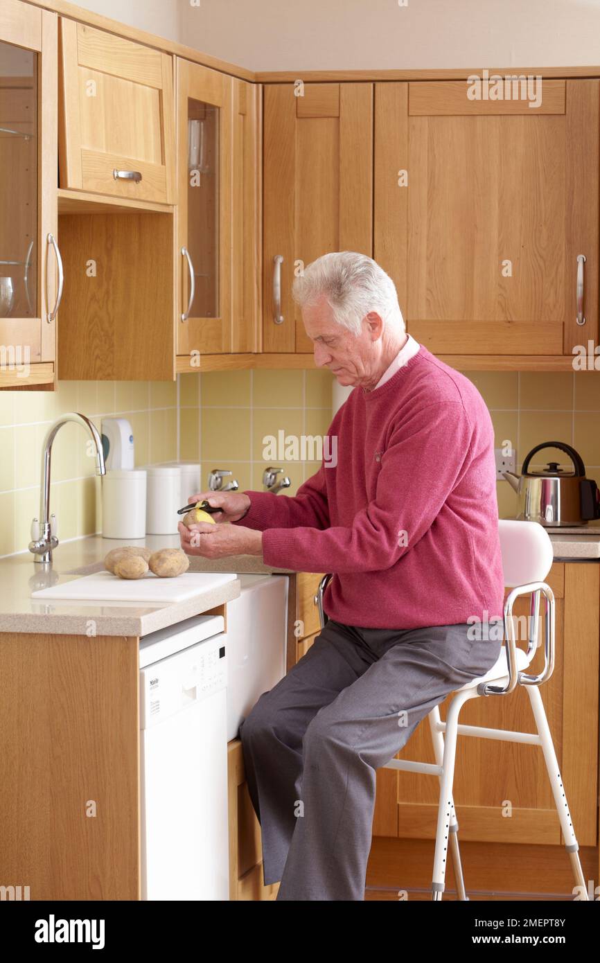 Mann sitzt auf einem Hochstuhl an der Küchenarbeitsfläche, um Kartoffeln zu schälen Stockfoto