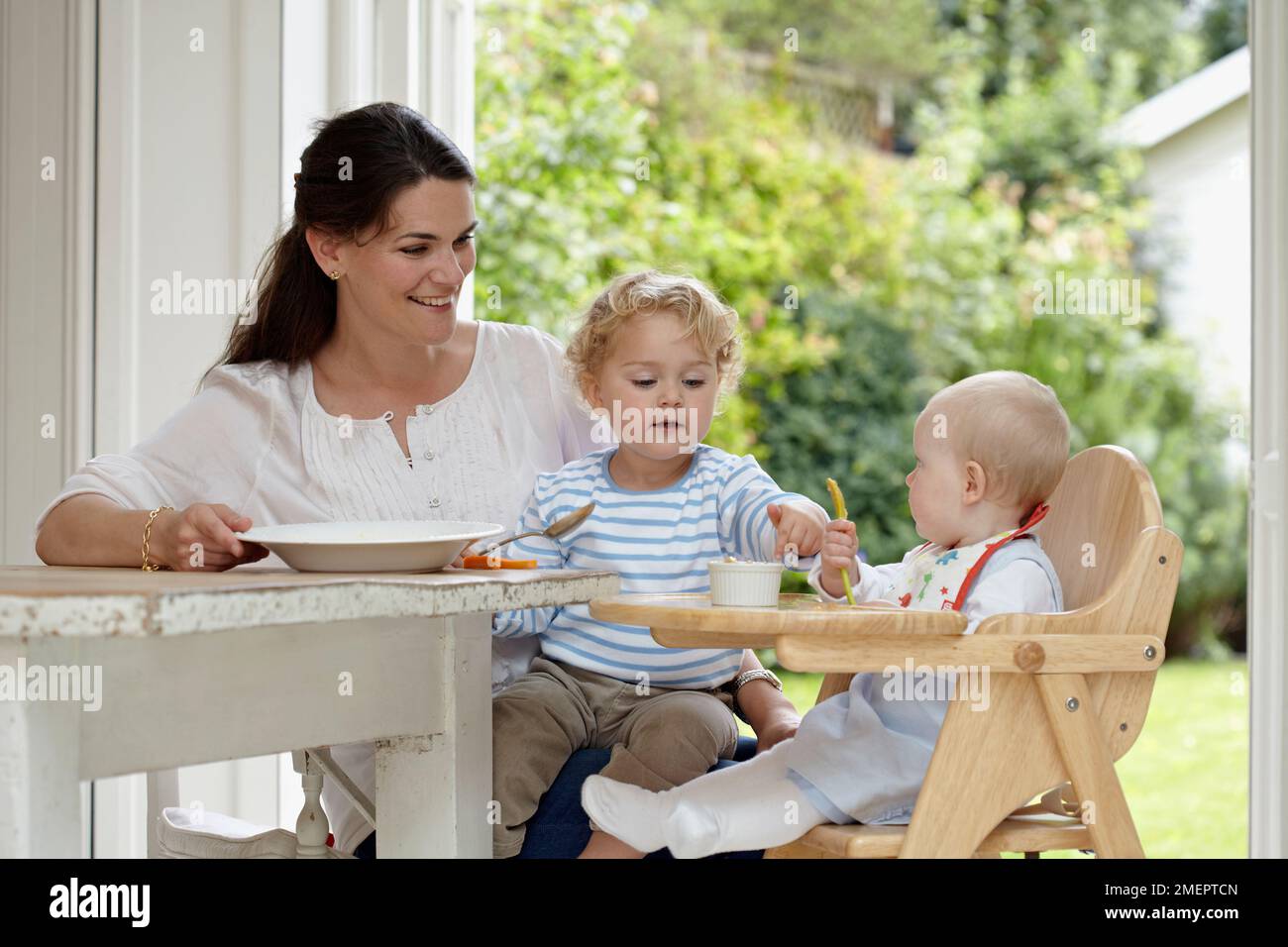 Frau sitzt am Tisch mit Kleinkind auf dem Schoß und Baby im Hochstuhl, 2 Jahre, 4 Monate und 9 Monate Stockfoto