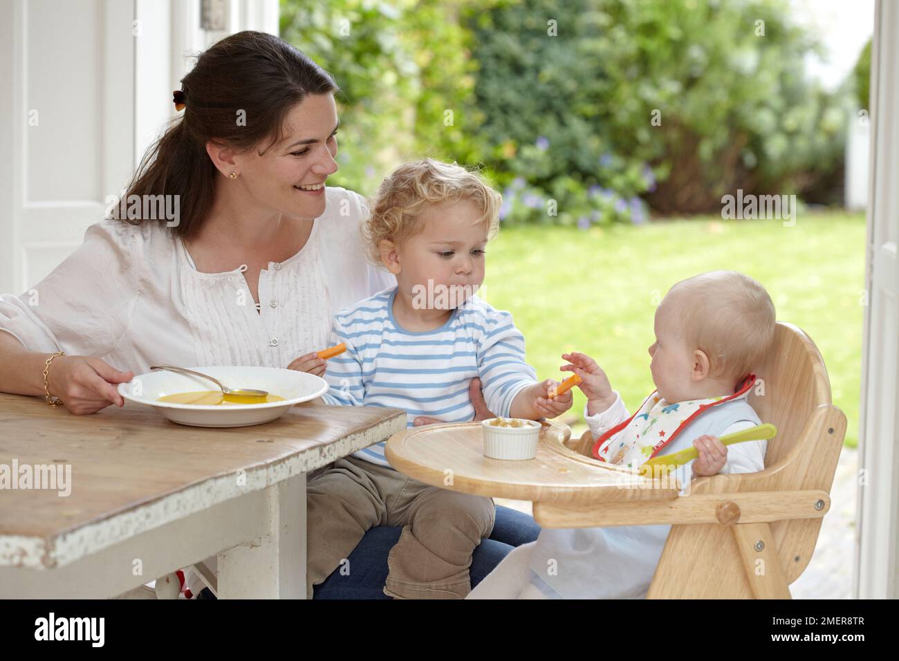Frau sitzt am Tisch mit einem Kleinkind (2 Jahre, 4 Monate) auf dem Knie und Baby (9 Monate) im Hochstuhl Stockfoto