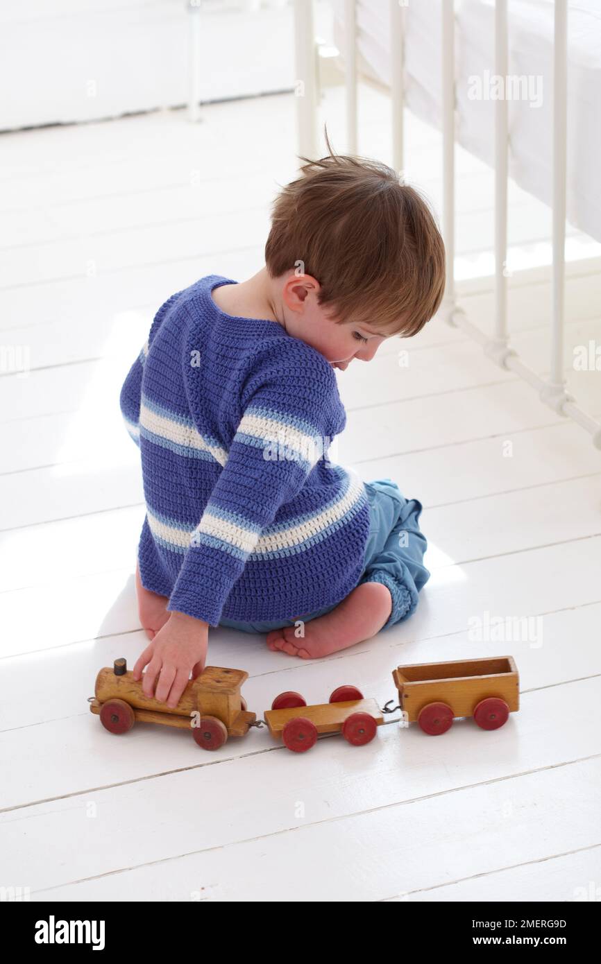 Ein Junge mit gestricktem Pullover kniet auf dem Boden und schiebt ein Spielzeug aus Holz, 2,5 Jahre Stockfoto