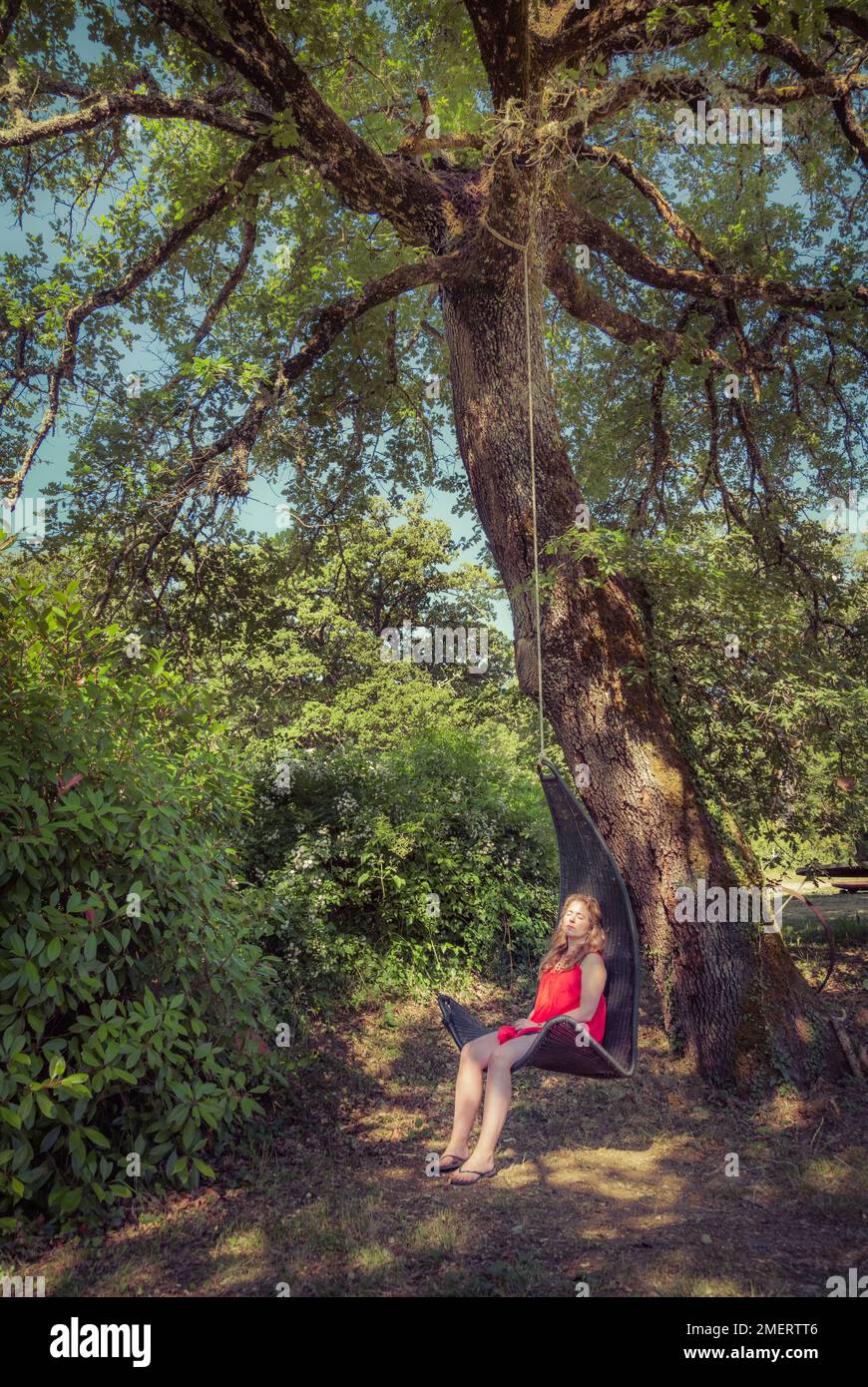 Eine junge Frau in rotem Kleid, die sich in der Schaukel entspannt, hängt an einem großen Baum in der Toskana, Italien. Stockfoto