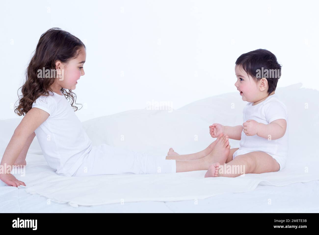 Kleines Mädchen (40 Wochen), das auf dem Bett sitzt und seiner Schwester (3,5 Jahre) gegenübersteht, die sich die Beine zu ihr streckt Stockfoto