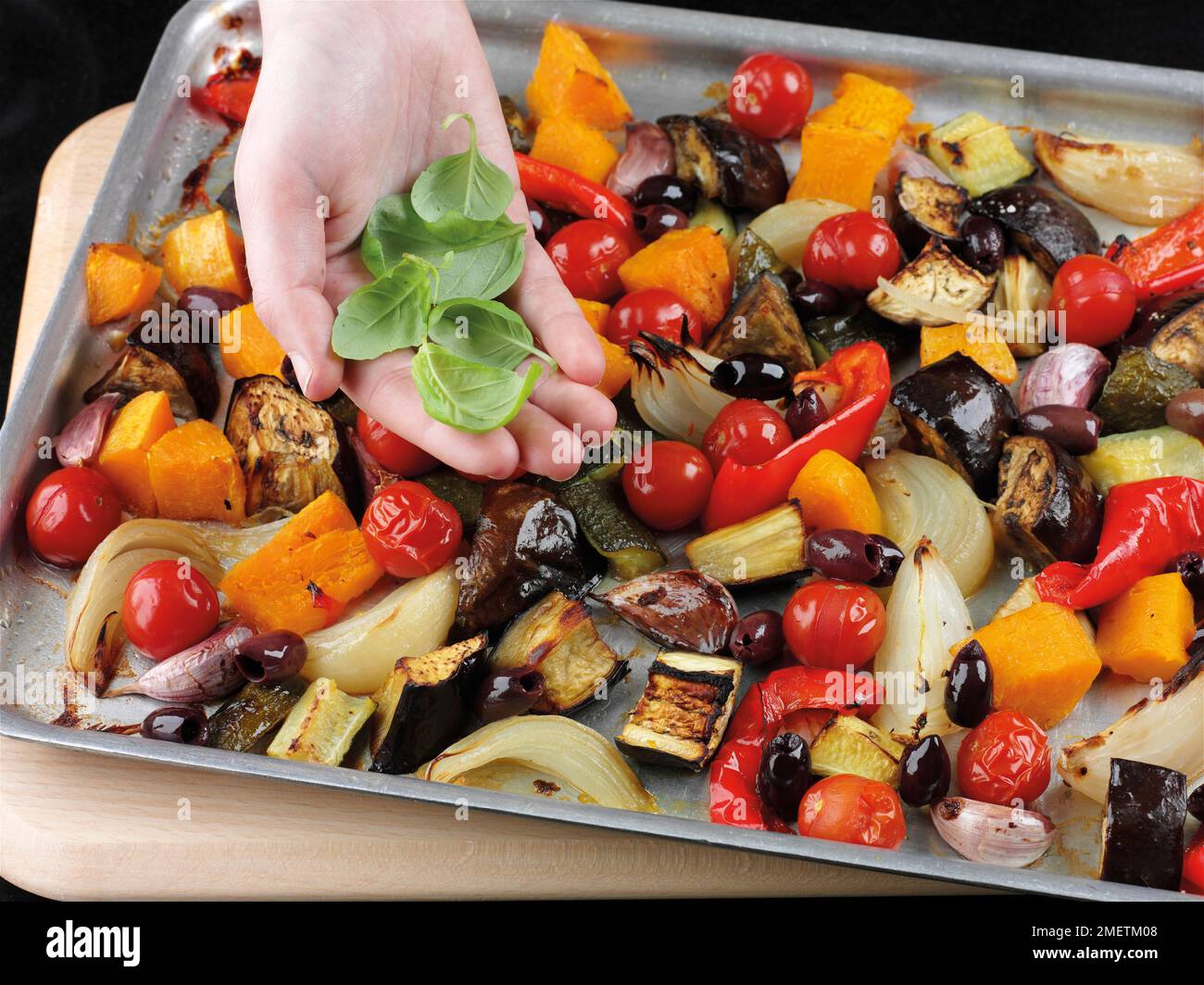 Zugabe von Oliven und frischen Basilikumblättern zu geröstetem Gemüse (Zucchini, Auberginen, Zwiebeln, Paprika, Kirschtomaten, butternusskürbis, Knoblauch) Stockfoto