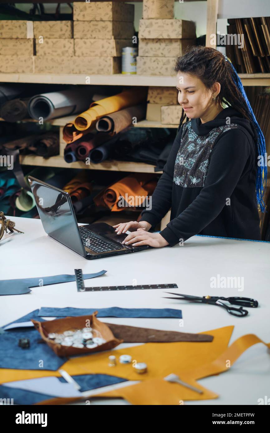 Online-Bestellung von Schneiderin. Eine junge Frau, die in der Schneiderei einen Laptop-Bildschirm ansieht. Modedesigner, Schneider Stockfoto