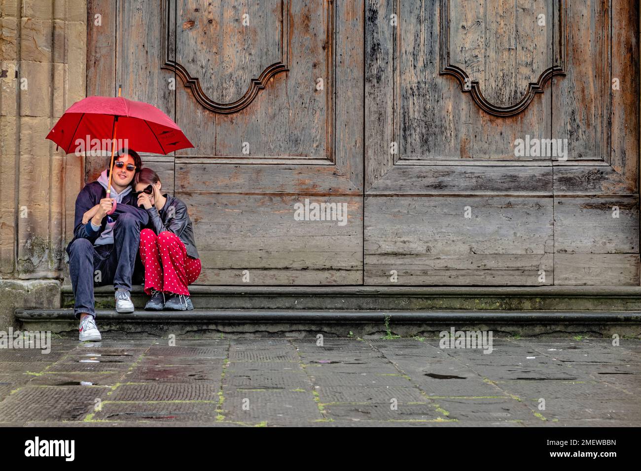 Ein junges Paar, das vor der Tür unter einem roten Schirm sitzt, in Florenz, Italien. Stockfoto