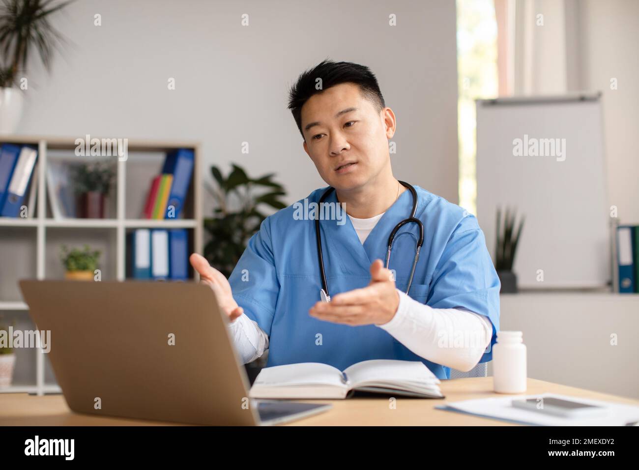 Seriöser, reifer chinesischer Therapeut in Uniform, Computer anschauen, sprechen, Gesten am Tisch in der Klinik Stockfoto