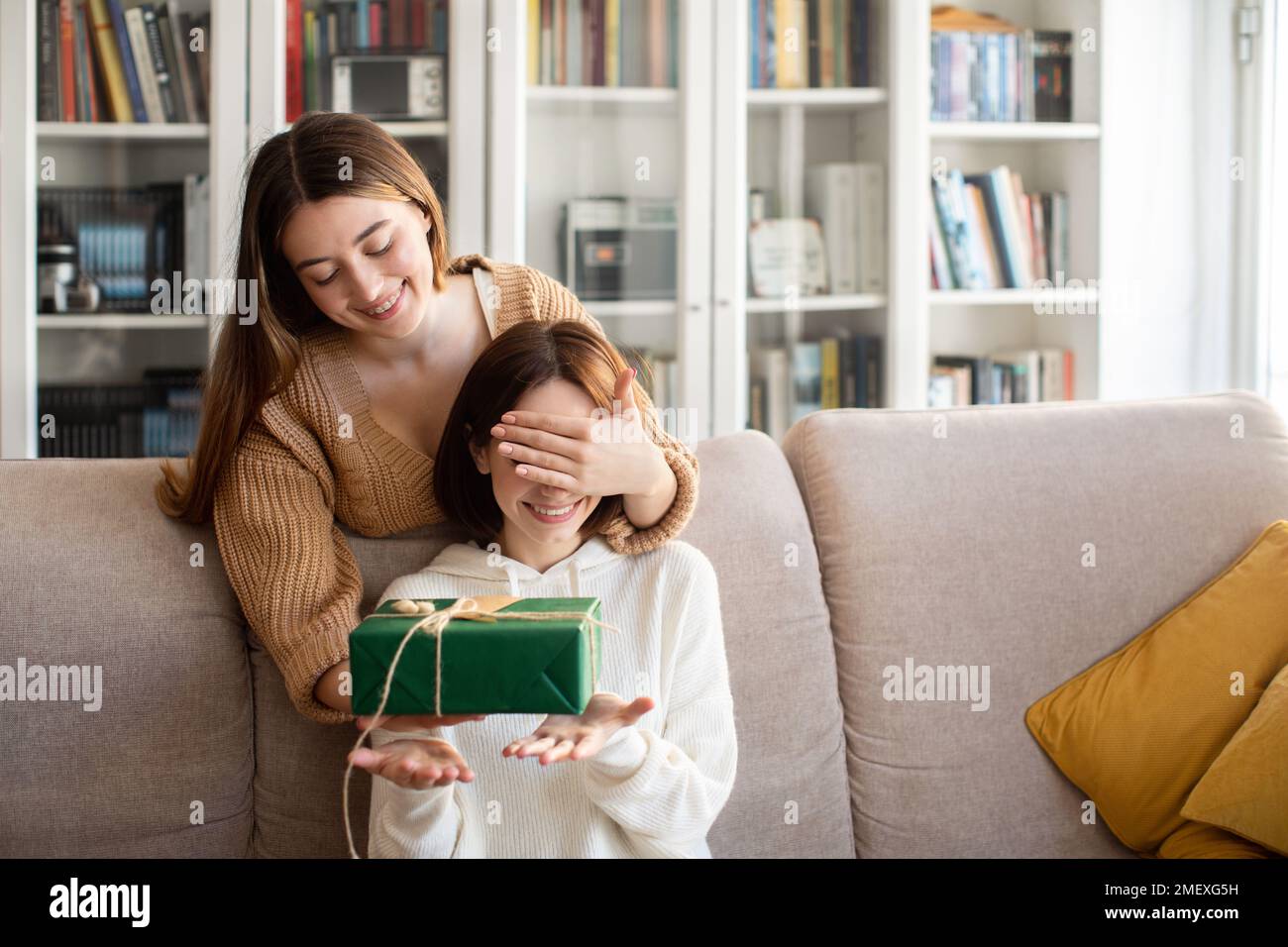 Die fröhliche europäische Millennials-Dame schließt die Augen vor ihrem Freund und schenkt ihr eine Geschenkbox im Wohnzimmer Stockfoto