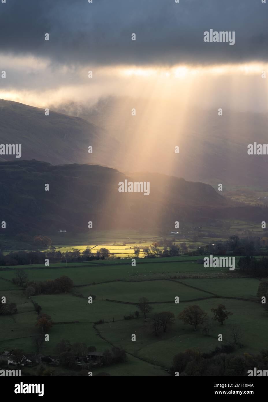 Lichtstrahlen, die an einem Sommermorgen durch dunkle und dramatische Sturmwolken über grünen Feldern in britischer Landschaft scheinen. Lake District, Großbritannien. Stockfoto