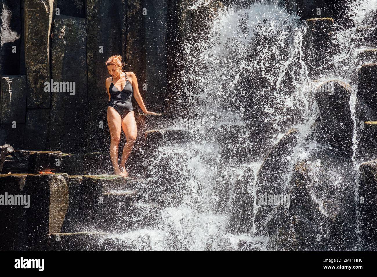 Weiße Frau in schwarzem Badeanzug erfrischt sich unter fallenden Wasserströmen, die über schwarze Vulkansteinkaskaden fließen. Wasserfall Rochester Falls - Popul Stockfoto
