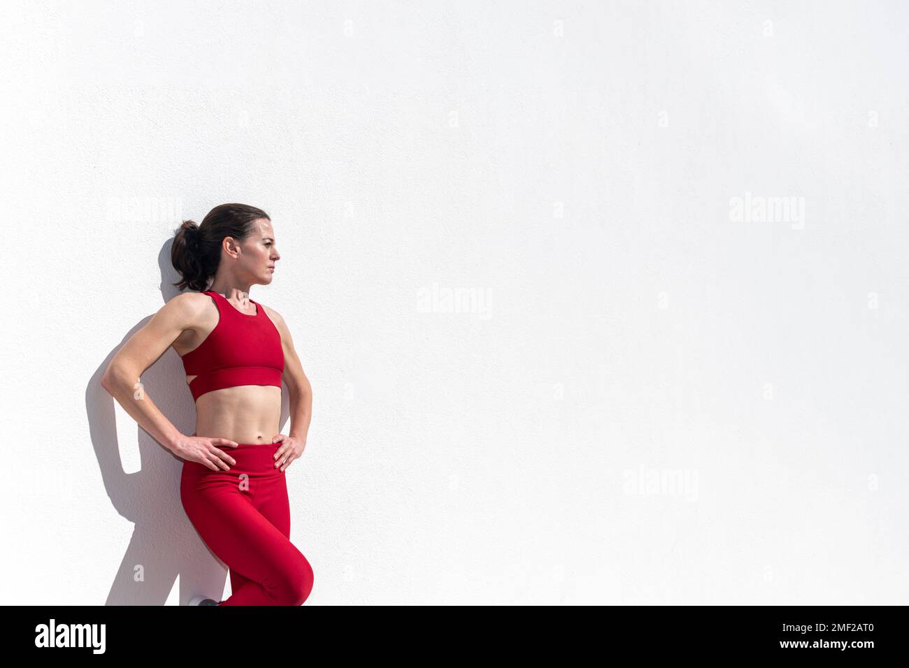 Sportliche Frau mit Händen an der Hüfte, die sich in der Sonne gegen eine weiße Wand lehnt. Stockfoto