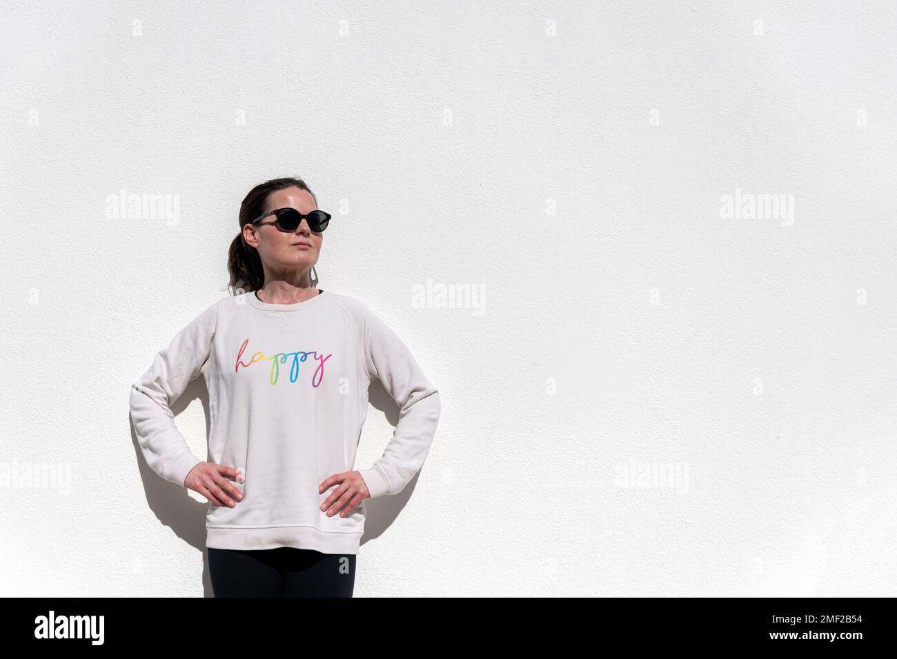 Eine Frau, die an einer weißen Wand steht und Hände auf der Hüfte hat. Sie trägt ein Sweatshirt mit dem „Happy“-Slogan. Stockfoto