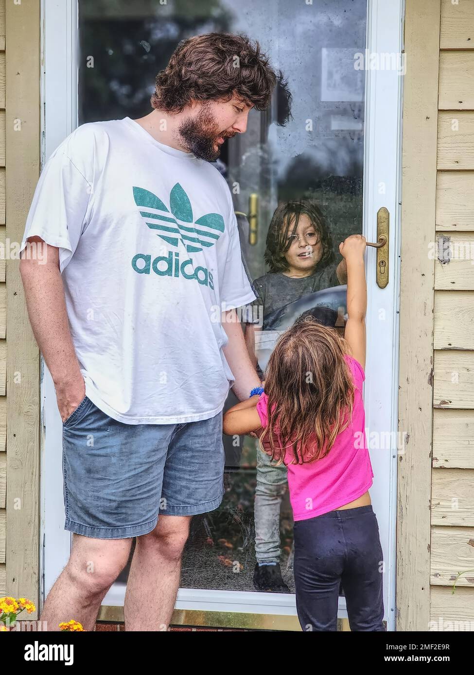 Ein junger Mann mit einem kleinen Mädchen draußen, in der Nähe einer Tür mit einem Jungen drinnen Stockfoto