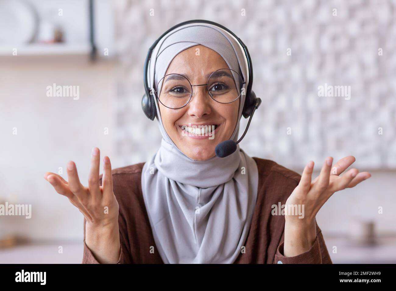 Hidschab-Frau, die zu Hause arbeitet, muslimische Frau mit einem lächelnden Headset für Videoanrufe, die in die Kamera schaut, mit dem Kunden online in der Küche spricht, Webcam-Ansicht pov. Stockfoto